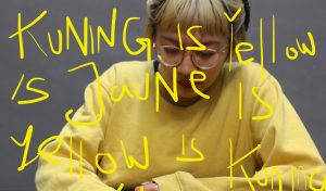 Yellow text written 'Kuning is Yellow is Jaune is Yellow is tuning is Yellow' in the colour yellow with the artist wearing a yellow sweater in the grey background.