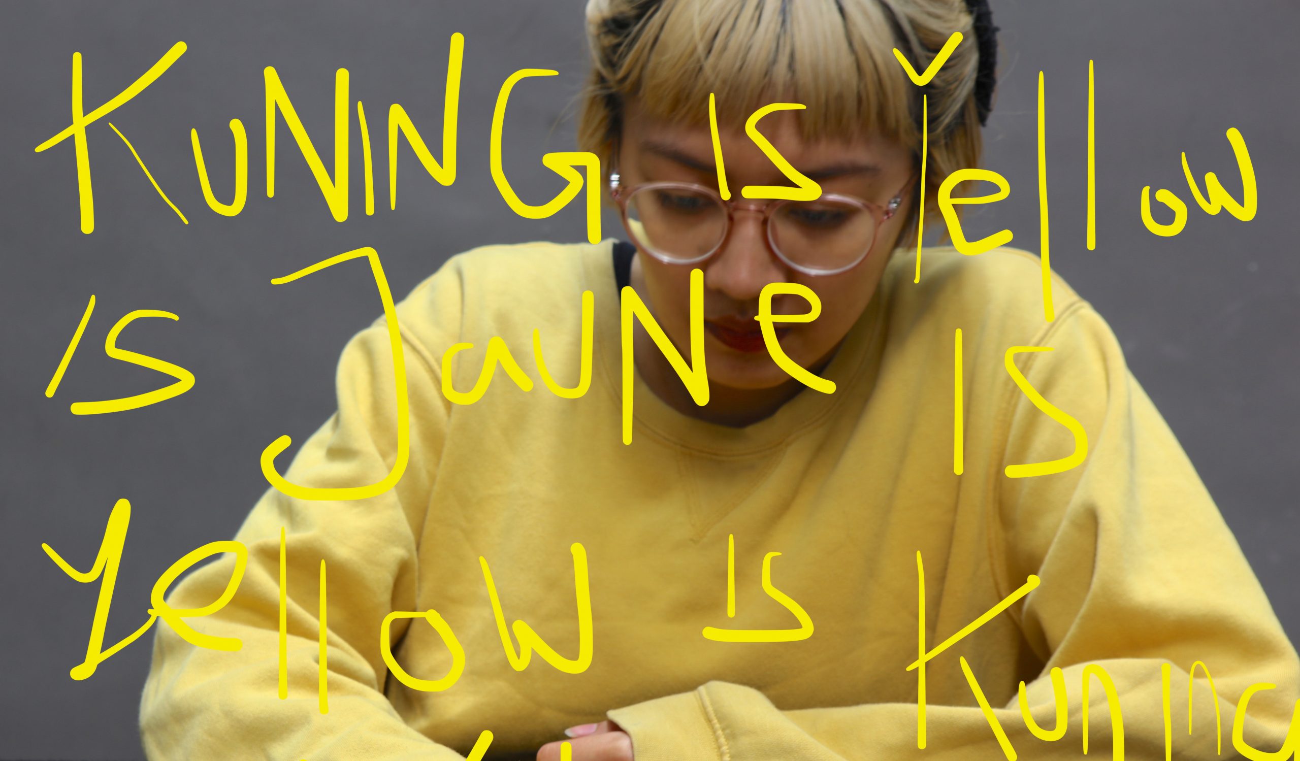 Yellow text written 'Kuning is Yellow is Jaune is Yellow is tuning is Yellow' in the colour yellow with the artist wearing a yellow sweater in the grey background.