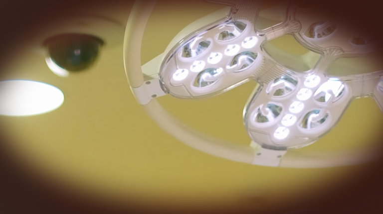 A screenshot from a film, there is the ceiling of a hospital room with a surgical light, there is a dark circle around the image creating a point of view effect, as though the viewer is a patient looking up at a hospital light.