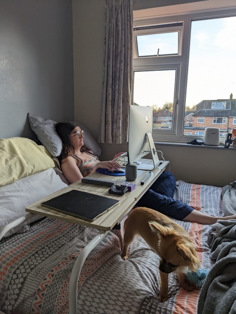 A photo of Charlie in a bed with a bed desk over her and working on a computer which is on top of the desk. There is a window next to the bed which gives a view of other houses and there is a small chihuahua dog next to her legs.