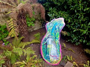 Photograph of my painting in the garden. The image is painted on a wooden board, it has a pink edge all the way around it, and has figures emerging from a green landscape.