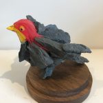 A bird hybrid sculpture - the head of a lifelike red bird model, attached to a group of dark grey rocks that have been glued together to form the bird’s body - two wings flapped out, a body and a tail.