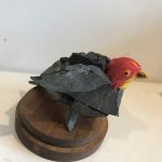 A bird hybrid sculpture - the head of a lifelike red bird model, attached to a group of dark grey rocks that have been glued together to form the bird’s body - two wings flapped out, a body and a tail.