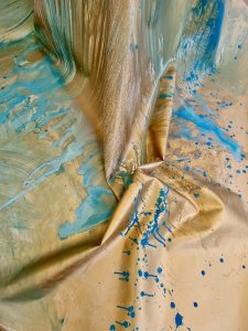 photograph of a close up of the surface of a canvas. There are drips of blue paint merging into the golden paint.