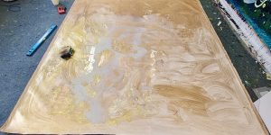 Photograph of a canvas on the floor with a golden wash of acrylic paint.