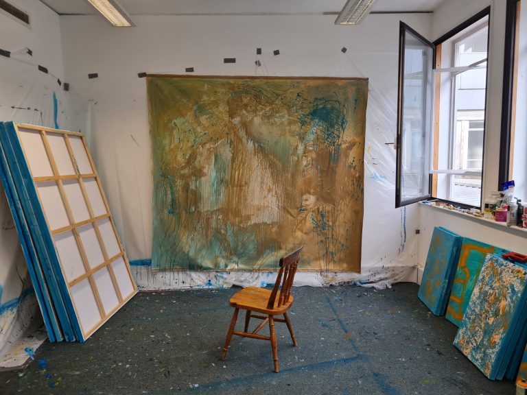 Studio shot of Latifah's studio- chair in fron of the painting that is against wall and stretched canvases on the left and right walls with a window on the right wall