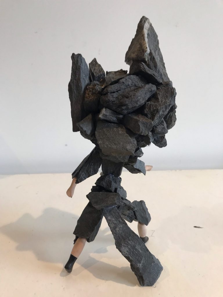 A girl hybrid - group of dark grey rocks are attached as the head of the girl, which is attached to a group of dark grey rocks that have been glued together to form the girls body - two hands and feet are not made of rocks