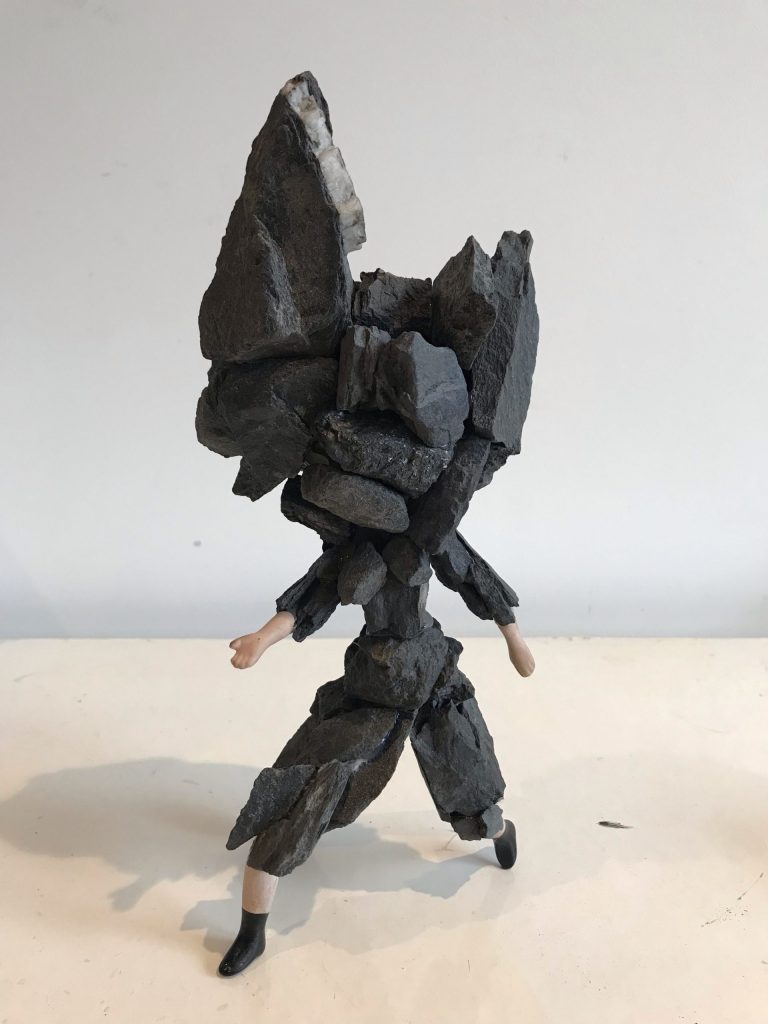 A girl hybrid - group of dark grey rocks are attached as the head of the girl, which is attached to a group of dark grey rocks that have been glued together to form the girls body - two hands and feet are not made of rocks
