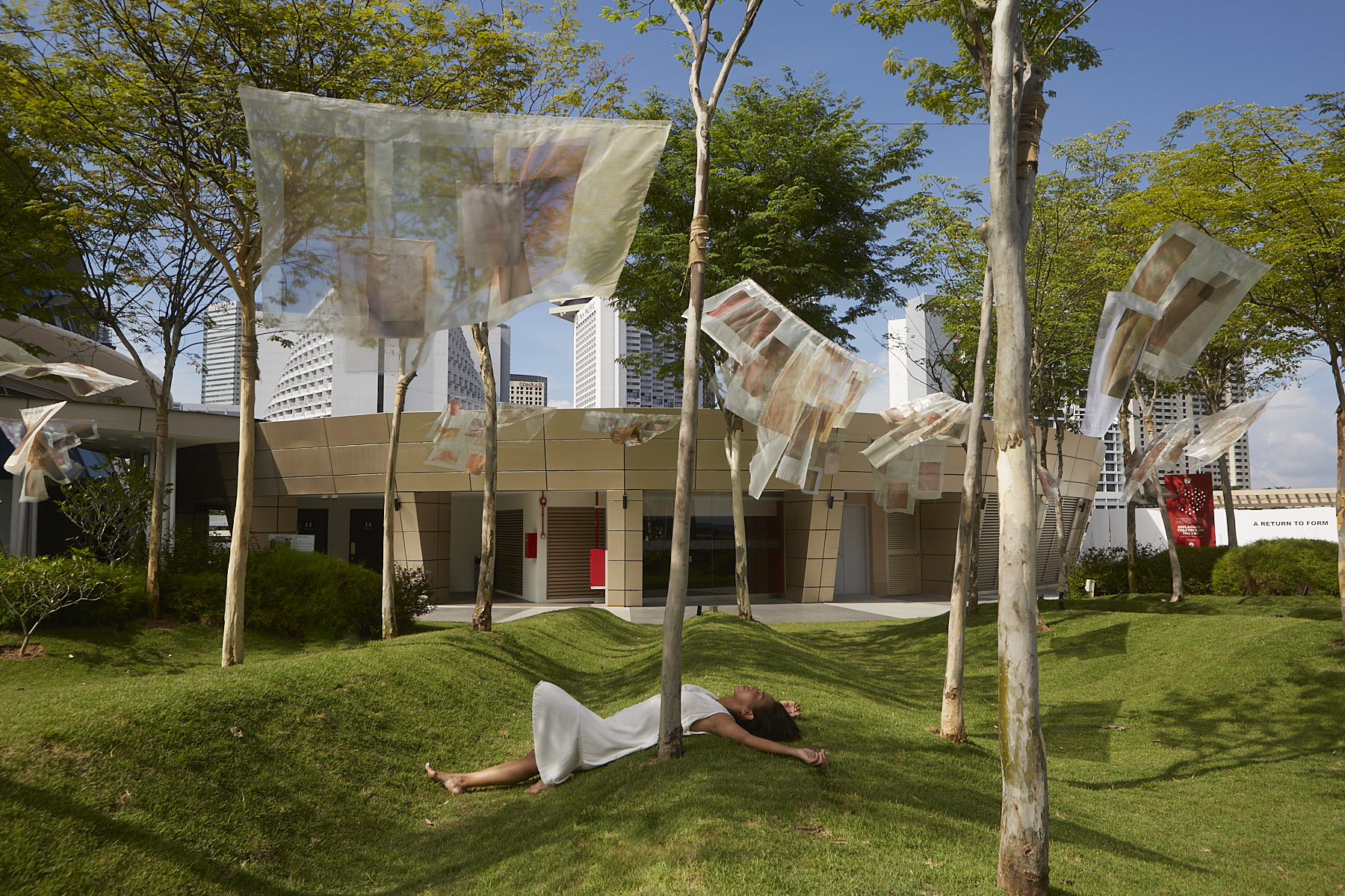 A view of an Asian women in a white dress, laying down on the grass below the trees and under the see through fabric installation being blown by the breeze