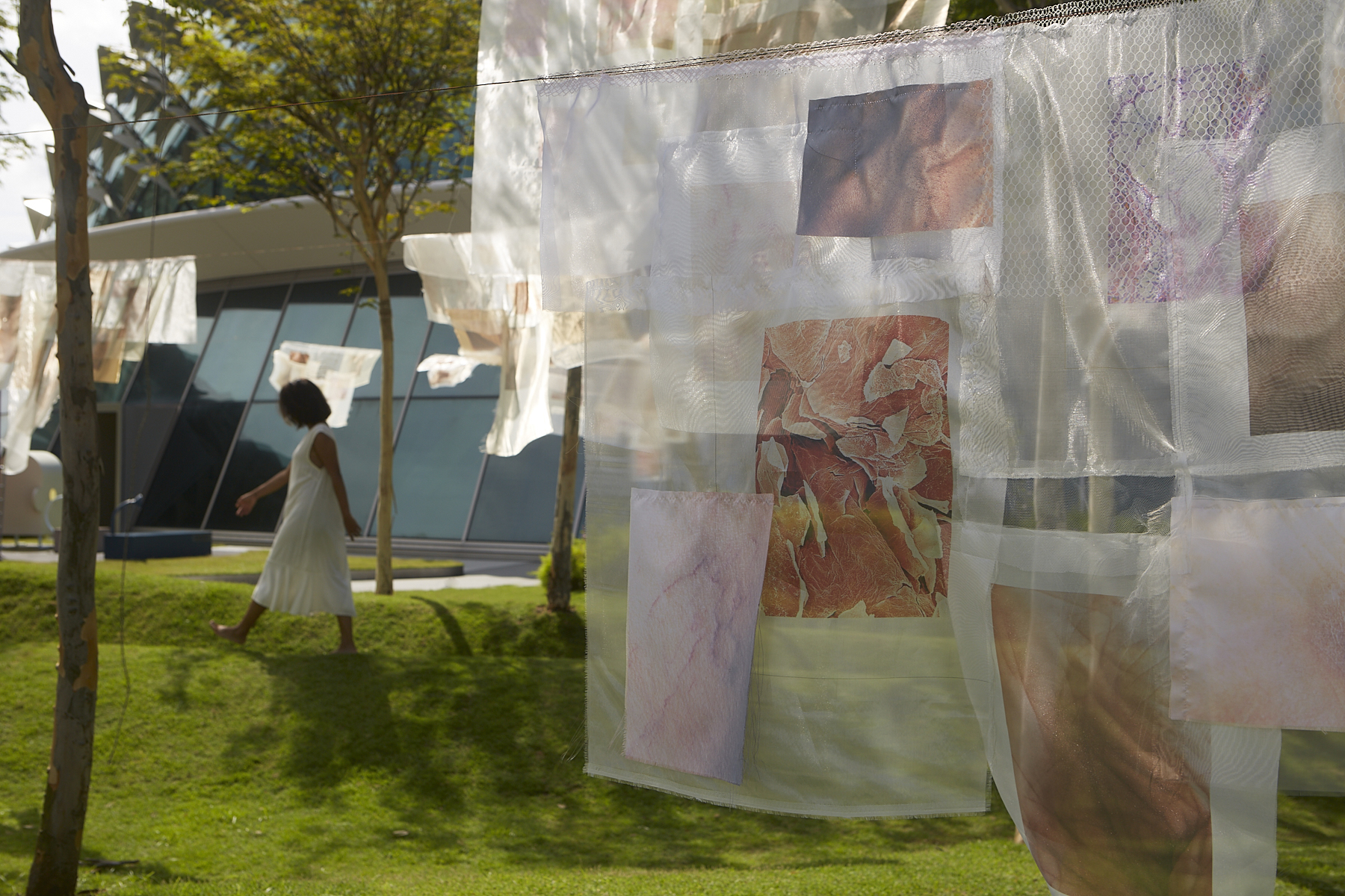A view of an Asian woman in a white dress walking the background, with the white see-through fabric installation in the foreground hanging down revealing the neutral-coloured images