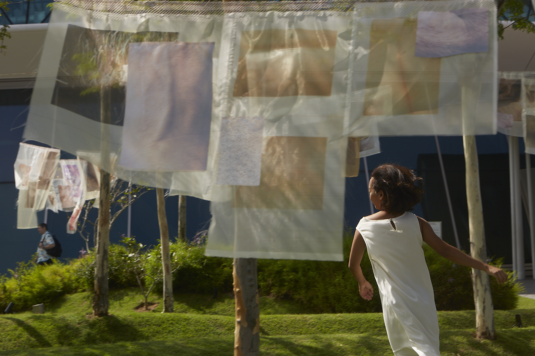A view of the back of an Asian woman in a white dress in the background - with the white see-through fabric installation in the foreground hanging down revealing the neutral-coloured images of human skin