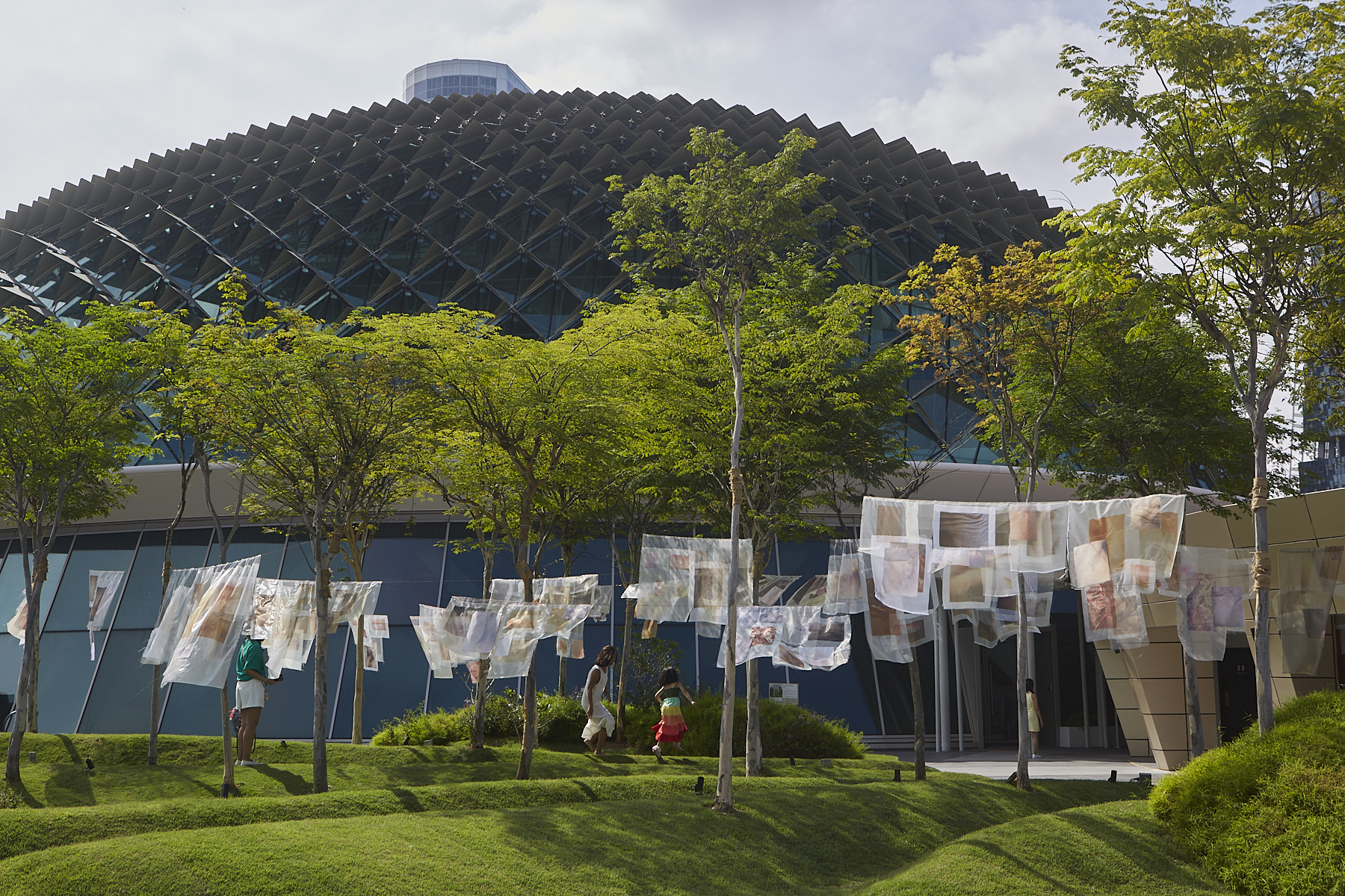A view transparent neutral- coloured fabric installation hung across trees in front of a domed building - with Asian women and girl in the background