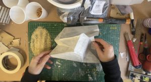 Hand holding a grey shaped structural sculpture on a working studio table full of working tools