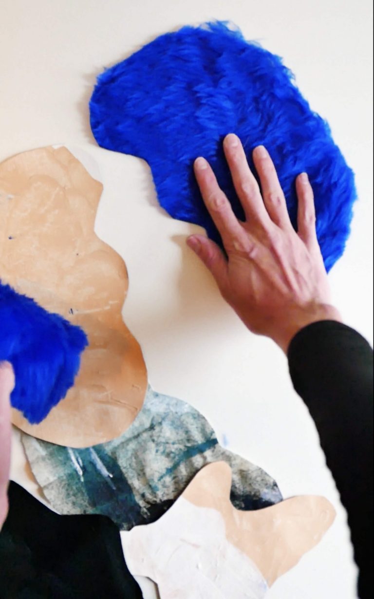 Hand touching against a blue furry organic shape cut out against a white wall
