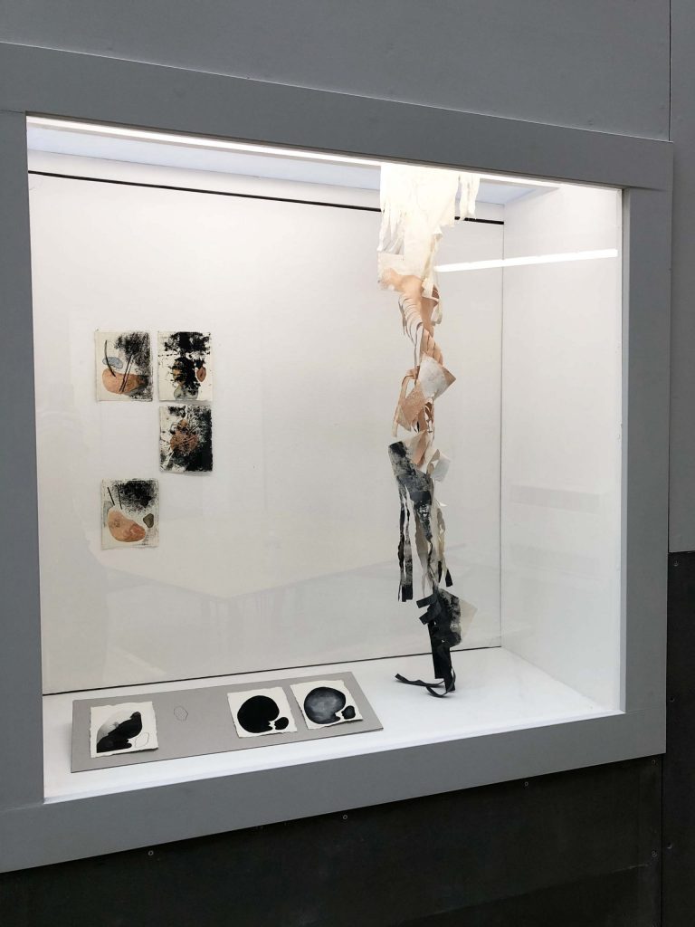 Black, white and skin colour abstract paintings on A4 size papers against wall and on the floor display, one long one hanging from the top of a cube display box
