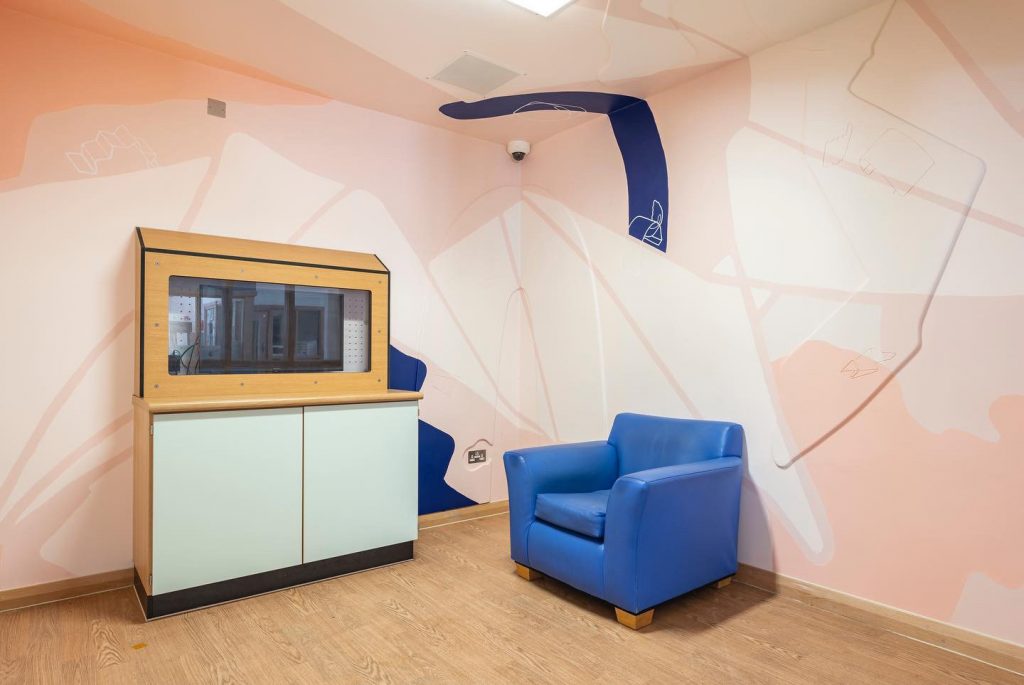 A corner view of a 4-colour mural with MDF reliefs, to the left is a tall wooden TV unit and on the right a blue armchair