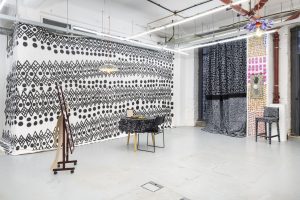 Installation including black and white painted wallpaper on the left hand side, white painted patterned chairs and tables at the centre, brown 4 foot 5 inch sized slim mirror, with a slightly shorter dancing female figure leaning against it. Towards the back, long, draping black and white curtains hang in front of the 10 foot tall window, and the ends of the curtains drape slightly across the ground in front of it. Right next to this is a four part multicoloured digital drawing palboard wallpaper narrow installation, between the curtains and the entrance. On top of this plaboard hangs a small painting. Lastly, in front of the narrow wallpaper, stands another black chair covered in hand drawn patterns.