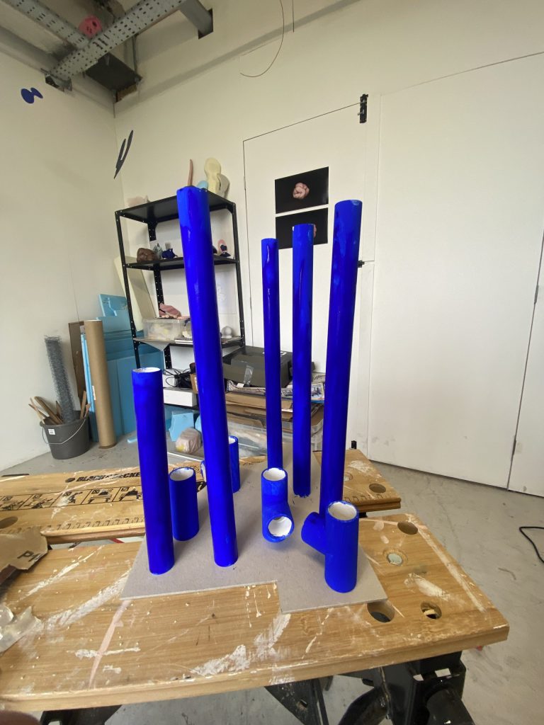 A series of plastic tubes and T connectors painted in cobalt blue drying on a workbench in the middle of Naomi’s studio