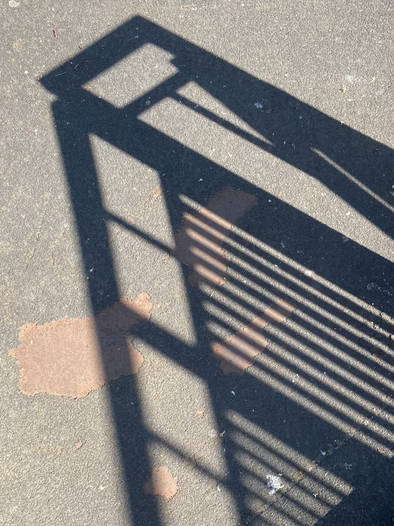 A shadow of the corner of bridge railings cast on mottled tarmac floor