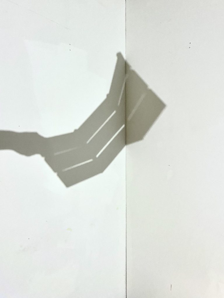 A shadow of a hand holding a 3 side crate like structure against a white wall