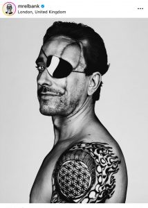 black and white photograph of a side profile of a man who looks striking, he has a black eye patch covering his right eye with a large visible scar above the eye in the shape of an arch. he has a flaming tattoo on his shoulder. He has short dark hair which is slicked back. he has stubble and a moustache.