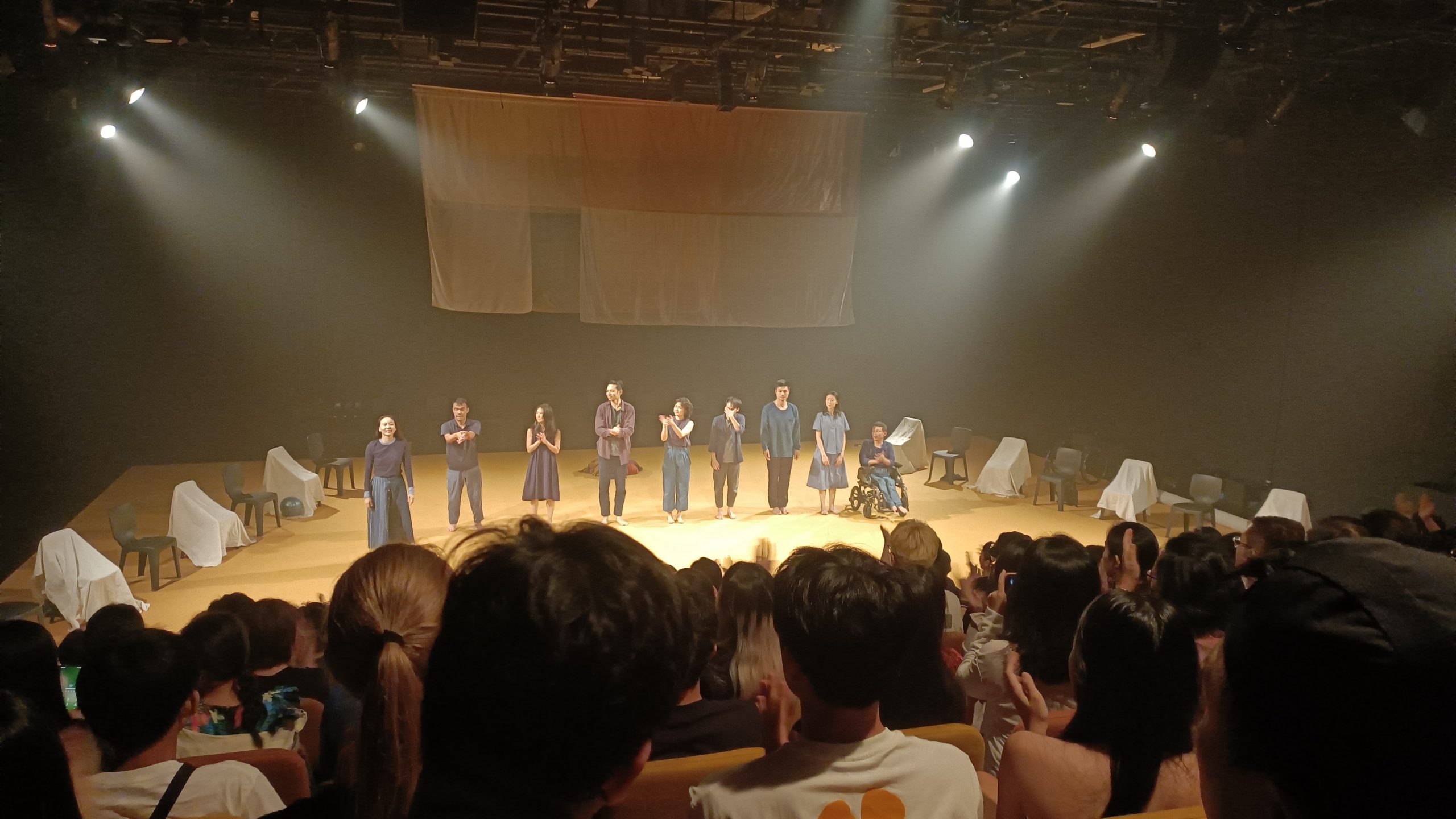 In a curtain call, the theatre is warmly lit. Audiences on the seats and performers on the stage are clapping their hands.