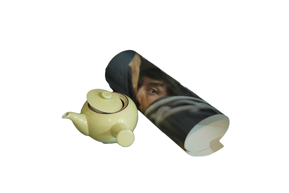 A yellow camomile colored teapot and a rolled poster, printed on the poster is a teenage boy